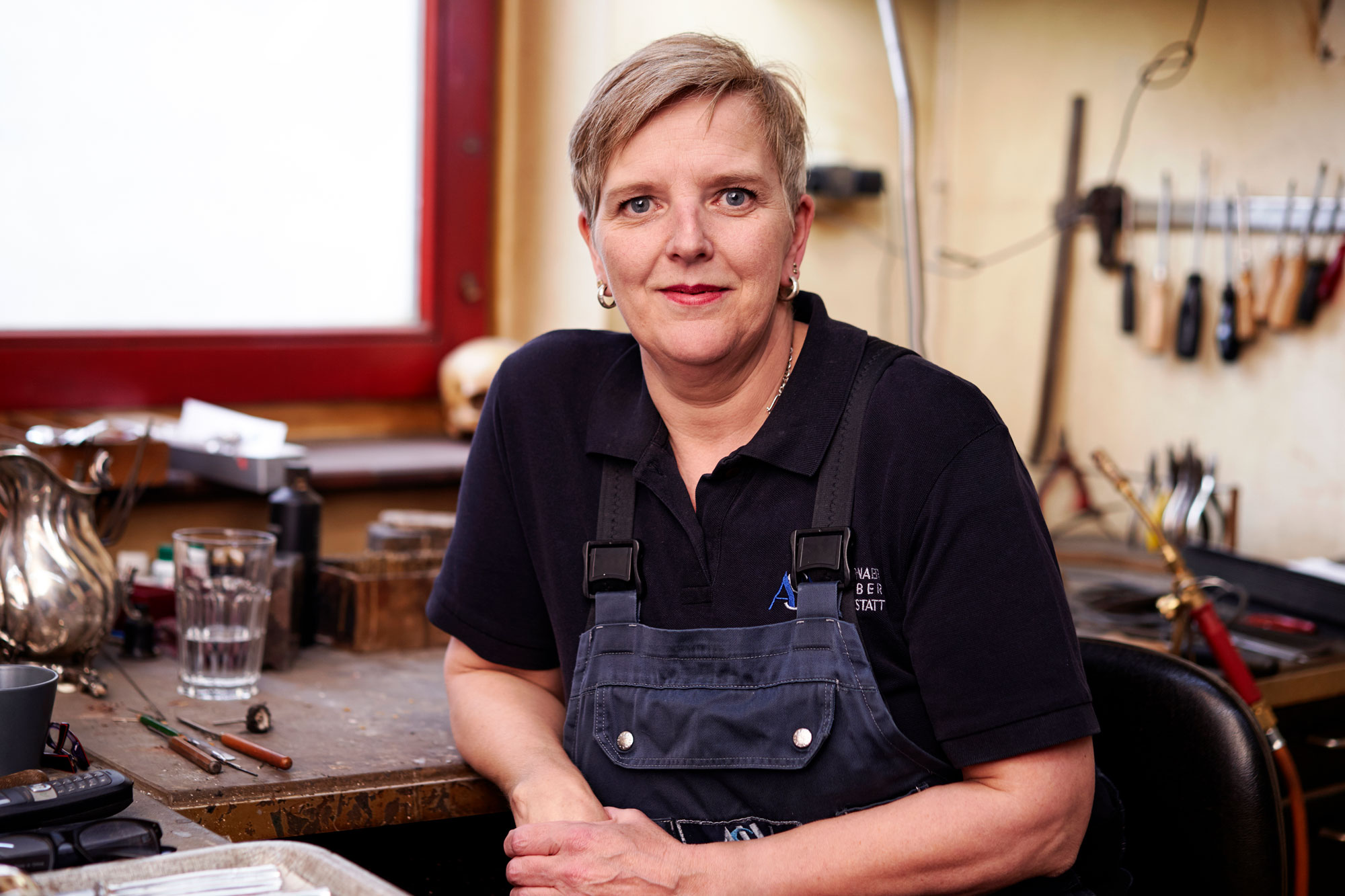 Eine blonde lächelnde Frau in einer Latzhose vor einer Schmiedebank